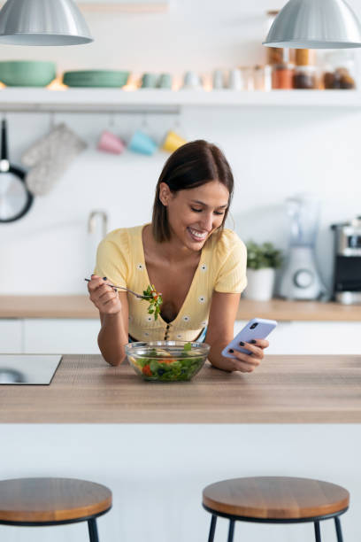 istock Beautiful young woman using her mobile phone while eating a salad in the kitchen at home. 1265293377