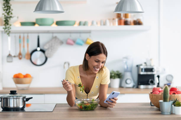 istock Beautiful young woman using her mobile phone while eating a salad in the kitchen at home. 1265294868