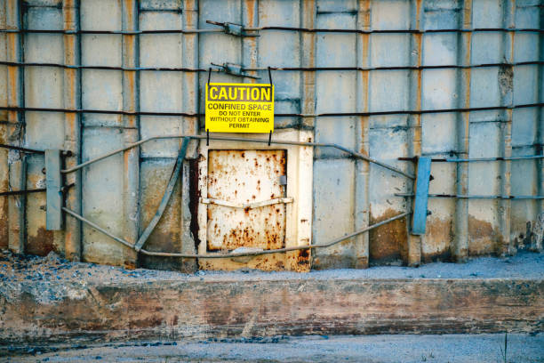 A bright yellow caution sign hangs over a small rusty metal door in a concrete wall. stock photo