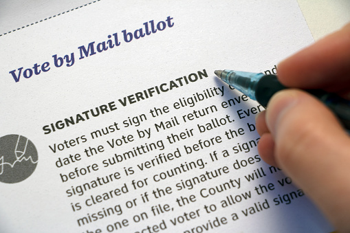 Closeup of a mail voter information paper, focused on 'Signature Verification' section and hand with pen hovering over it.