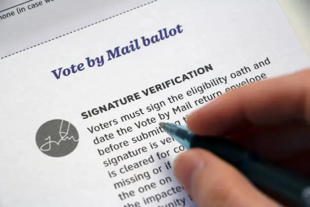 Closeup of a mail voter information paper, focused on 'Signature Verification' section and hand with pen hovering over it.
