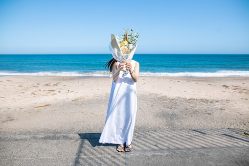 Young woman standing on beach and covering her face with bouquet