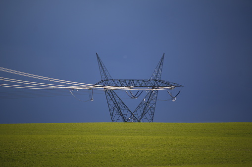 The tower and cables of an electrical power transmission line are set against ominous dark storm clouds with a yellow agricultural field in the foreground.
