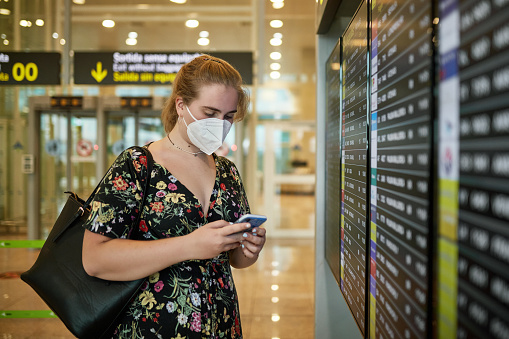 Partial view of Caucasian teenage girl in print dress and KN95 mask checking smart phone as she stands at airport arrival departure board in time of COVID-19.