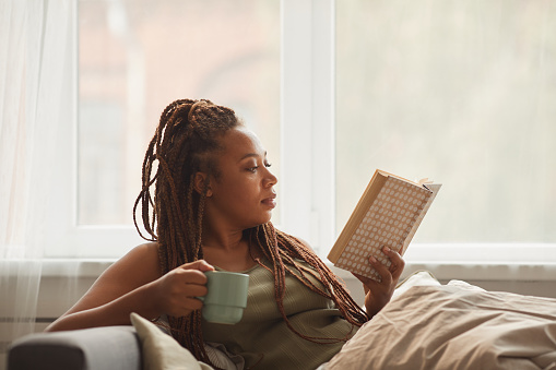 African young woman lying on sofa drinking coffee and reading an interesting book at home