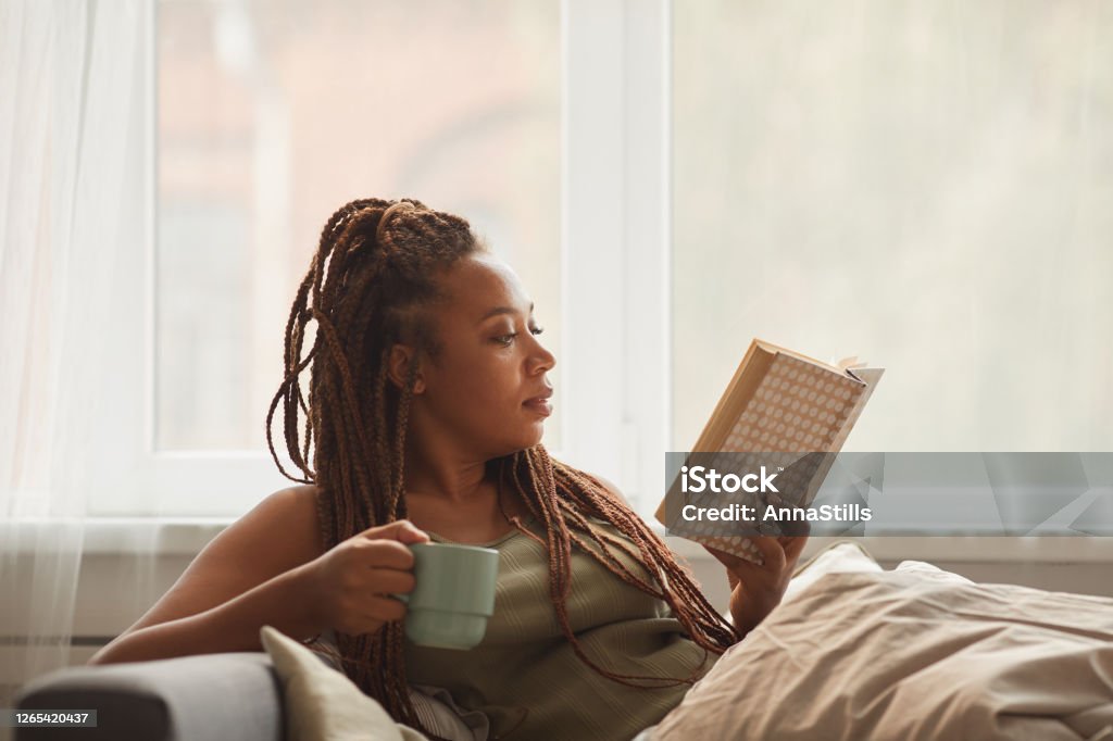 Woman reading a book African young woman lying on sofa drinking coffee and reading an interesting book at home Reading Stock Photo