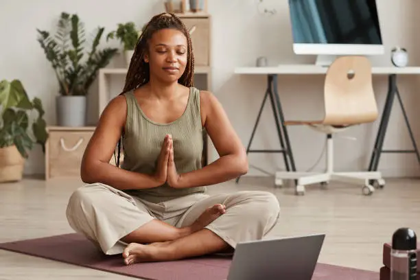 Young woman sitting in lotus position on exercise mat with her eyes closed and meditating in the living room