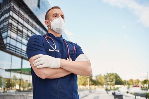 Medical doctor in uniform standing in front of a medical building, hospital, wearing N95 protective face mask and surgical gloves during coronavirus, covid-19 epidemic.