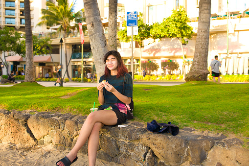 Biracial Asian Caucasian teenage girl tourist relaxing and sitting on stone wall in Waikiki, Honolulu, Hawaii at Kuhui beach eating ham musubi