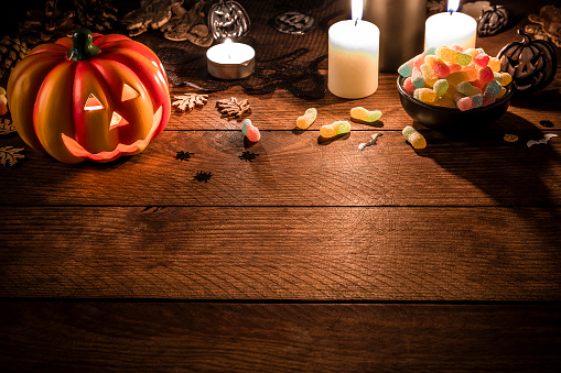 Halloween ornate: Front view of a ceramic Jack-o-Lantern pumpkin, some halloween ornate, some burning candles and a bowl full of gummy worms on a rustic wooden table. All the objects are at the top of the image leaving a useful copy space at the lower side on the wooden table. Low key DSLR photo taken with Canon EOS 6D Mark II and Canon EF 24-105 mm f/4L