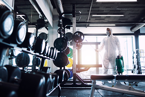 Desinfección y atención sanitaria en el gimnasio. Hombre con protección blanca traje de desinfección y equipos de fitness y pesas para dejar de propagar coronavirus altamente contagioso o COVID-19. photo
