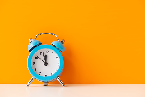 Blue alarm clock on white table in front of orange  wall. The clocks time is showing 5 to 12.