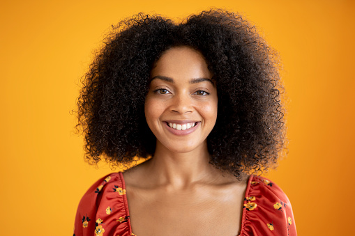 Front view of 23 year old woman with curly medium-length hair wearing casual print top with scooped neck and smiling at camera against orange background.