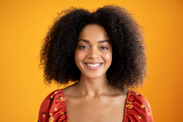 Headshot portrait of cheerful young mixed race woman in summer top Front view of 23 year old woman with curly medium-length hair wearing casual print top with scooped neck and smiling at camera against orange background. mid length hair stock pictures, royalty-free photos & images