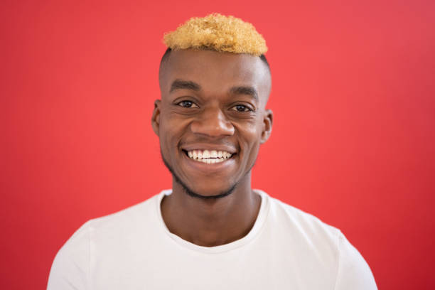 Portrait of casual 25 year old black man smiling at camera Indoor close-up of cheerful young black man with two-toned half-shaved hairstyle and facial hair wearing white t-shirt against red background. black guy with blonde hair stock pictures, royalty-free photos & images