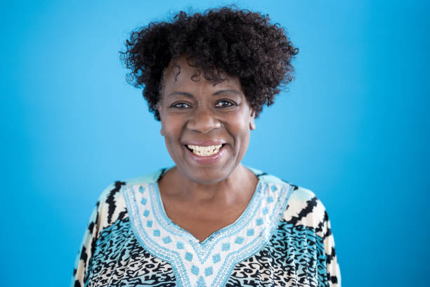 Studio portrait of 64 year old black woman smiling at camera Head and shoulders view of cheerful senior black woman with short curly hair wearing multi colored print top with border design against blue background. natural beauty people stock pictures, royalty-free photos & images