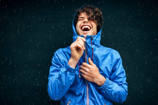 glücklicher mann lächelt breit, trägt blauen regenmantel während des regens draußen. schöne männchen in blauen regenmantel genießen den regen auf schwarze wand. fröhlicher mann hat fröhlichen ausdruck bei regnerischem wetter. - regenmantel stock-fotos und bilder