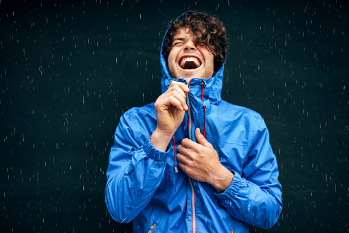 Happy man smiling broadly, wearing blue raincoat during the rain outside. Handsome male in blue raincoat enjoying the rain on black wall. Cheerful man has joyful expression in rainy weather.