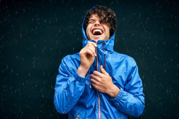 Happy man smiling broadly, wearing blue raincoat during the rain outside. Handsome male in blue raincoat enjoying the rain on black wall. Cheerful man has joyful expression in rainy weather.