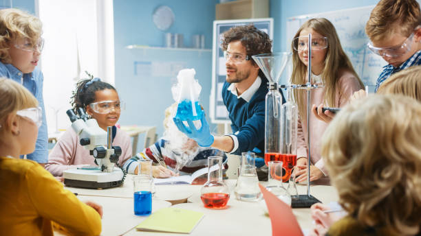 istock Elementary School Science Classroom: Enthusiastic Teacher Explains Chemistry to Diverse Group of Children, Shows them How to Mix Chemicals in Beakers. Children Use Digital Tablet Computers and Talk 1265582015