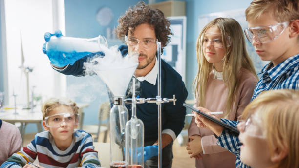istock Elementary School Science Classroom: Enthusiastic Teacher Explains Chemistry to Diverse Group of Children, Shows them How to Mix Chemicals in Beakers. Children Use Digital Tablet Computers and Talk 1265582025