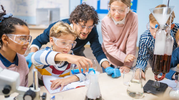 istock Elementary School Science Classroom: Enthusiastic Teacher Explains Chemistry to Diverse Group of Children, Little Boy Mixes Chemicals in Beakers. Children Learn with Interest 1265582034