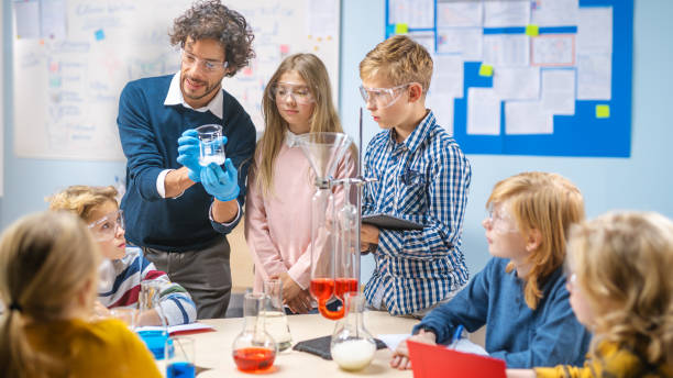 istock Elementary School Science Classroom: Enthusiastic Teacher Explains Chemistry to Diverse Group of Children, Shows them How to Mix Chemicals in Beakers. Children Look with Interest and Talk 1265582035