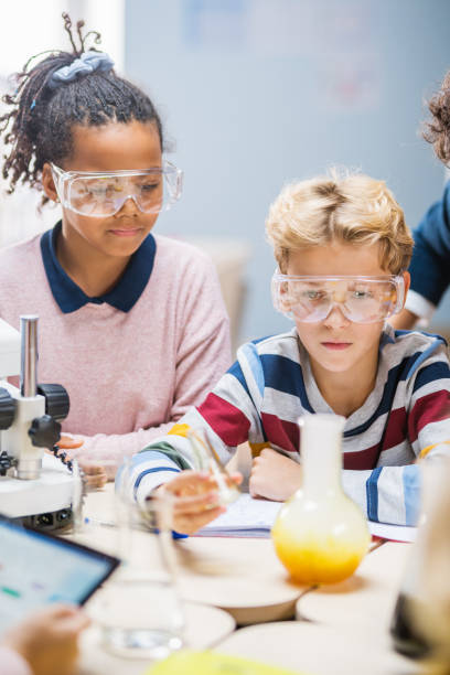 小学校の科学教室:安全メガネのかわいい男の子はビーカーの化学物質を混ぜます。子供たちは興味を持って化学科学を学ぶ - scientist research group of people analyzing ストックフォトと画像