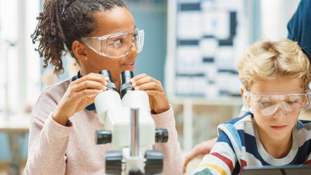 istock Elementary School Science Classroom: Cute Little Girl Looks Under Microscope, Boy Uses Digital Tablet Computer to Check Information on the Internet. Teacher Observes from Behind 1265582072