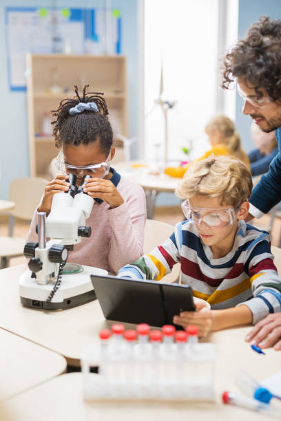 szkoła podstawowa science classroom: cute little girl wygląda pod mikroskopem, chłopiec używa cyfrowego tabletu do sprawdzania informacji w internecie. nauczyciel obserwuje od tyłu - science child laboratory education zdjęcia i obrazy z banku zdjęć