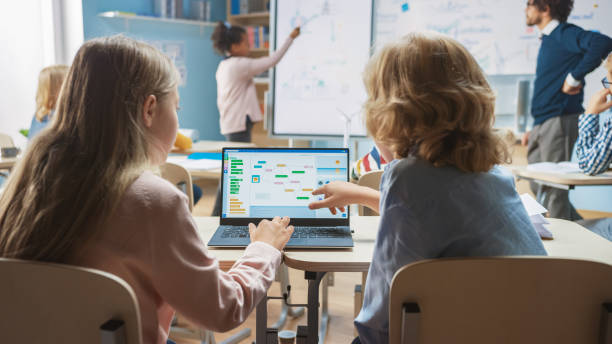 istock Elementary School Science Class: Over the Shoulder Little Boy and Girl Use Laptop with Screen Showing Programming Software. Physics Teacher Explains Lesson to a Diverse Class full of Smart Kids 1265582122