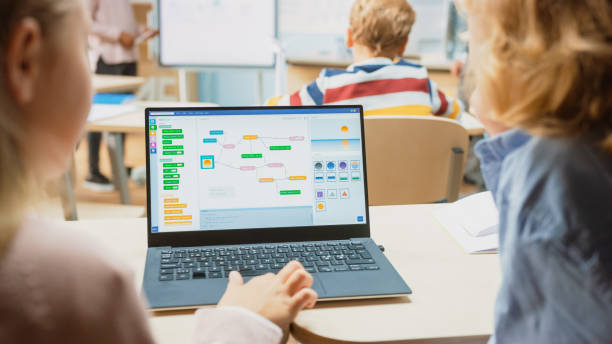 istock Elementary School Science Class: Over the Shoulder Little Boy and Girl Use Laptop with Screen Showing Programming Software. Physics Teacher Explains Lesson to a Diverse Class full of Smart Kids 1265582137