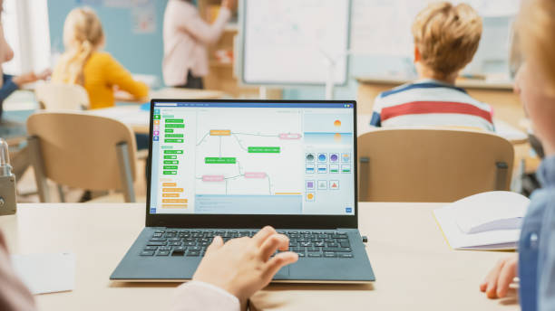 istock Elementary School Science Class: Over the Shoulder Little Boy and Girl Use Laptop with Screen Showing Programming Software. Physics Teacher Explains Lesson to a Diverse Class full of Smart Kids 1265582156