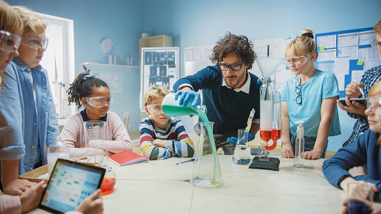 Elementary School Science / Chemistry Classroom: Teacher Shows Chemical Reaction Experiment to Group of Children. Mixing Chemicals in Beaker to get Reaction. Children Use Digital Tablet Computers
