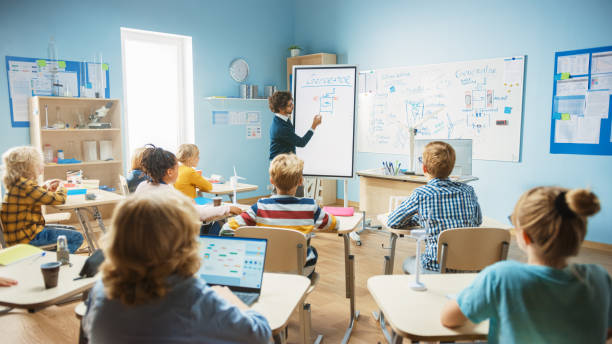 elementary school physics teacher uses interactive digital whiteboard to show to a classroom full of smart diverse children how generator works. science class with curious kids listening attentively - physics classroom teaching professor imagens e fotografias de stock