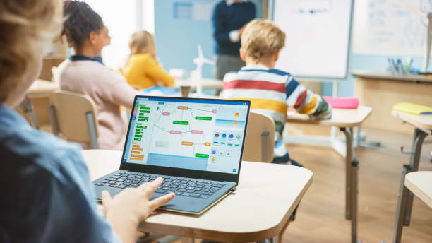istock Elementary School Science Class: Over the Shoulder Little Boy Uses Laptop with Screen Showing Programming Software. Physics Teacher Explains Lesson to a Diverse Class full of Smart Kids 1265582189