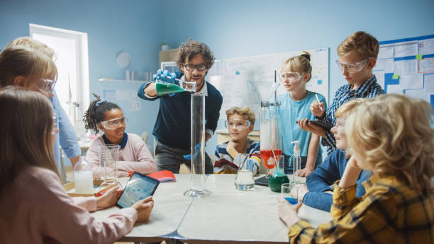 istock Elementary School Science / Chemistry Classroom: Teacher Shows Chemical Reaction Experiment to Group of Children. Mixing Chemicals in Beaker to get Reaction. Children Use Digital Tablet Computers 1265582197