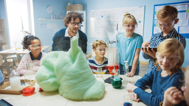 istock Elementary School Science / Chemistry Classroom: Enthusiastic Teacher Shows Funny Chemical Reaction Experiment to Group of Children. Mixing Chemicals in Beaker so they Shoot Foam (Elephant Toothpaste) 1265582204