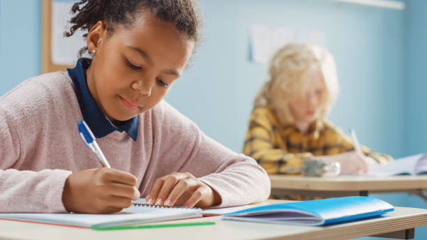 istock In Elementary School Classroom Brilliant Black Girl Writes in Exercise Notebook, Taking Test and Writing Exam. Junior Classroom with Group of Bright Children Working Diligently and Learning New Stuff 1265582222