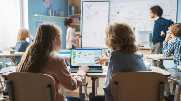 Elementary School Science Class: Over the Shoulder Little Boy and Girl Use Laptop with Screen Showing Programming Software. Physics Teacher Explains Lesson to a Diverse Class full of Smart Kids