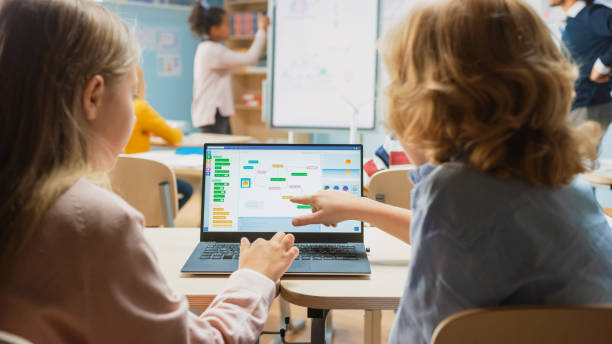 小学校の理科のクラス:肩の上に小さな男の子と女の子は、プログラミングソフトウェアを示す画面とラップトップを使用しています。物理学の先生は、スマートキッズでいっぱいの多様なク - elementary student little boys classroom child ストックフォトと画像