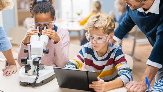 Elementary School Science Classroom: Cute Little Girl Looks Under Microscope, Boy Uses Digital Tablet Computer to Check Information on the Internet, while Enthusiastic Teacher Helps Boy