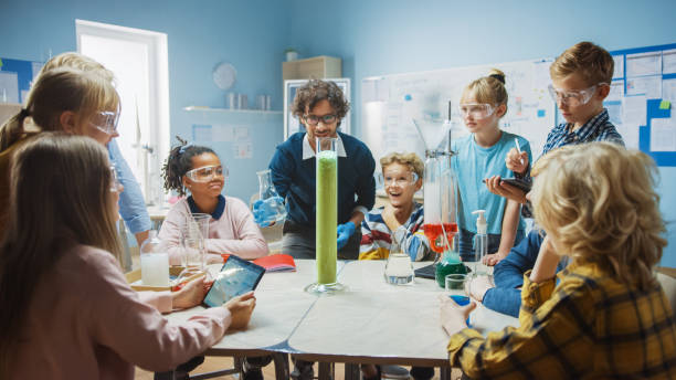 istock Elementary School Science / Chemistry Classroom: Teacher Shows Chemical Reaction Experiment to Group of Children. Mixing Chemicals in Beaker to get Reaction. Children Use Digital Tablet Computers 1265582277