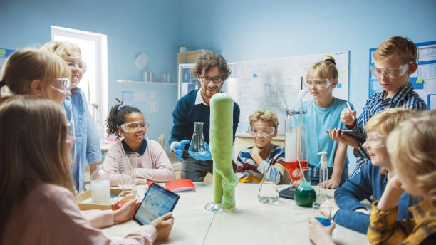 istock Elementary School Science / Chemistry Classroom: Enthusiastic Teacher Shows Funny Chemical Reaction Experiment to Group of Children. Mixing Chemicals in Beaker so they Shoot Foam (Elephant Toothpaste) 1265582283
