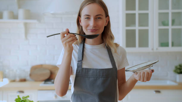 キッチンのストーブでスープを調理するかわいい女性。スプーンでスープを味わう若い女性 - tasting women eating expressing positivity ストックフォトと画像