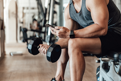 Close up shot of a young man lifting dumbbell while using smartphone. Bodybuilding motivation and determination.