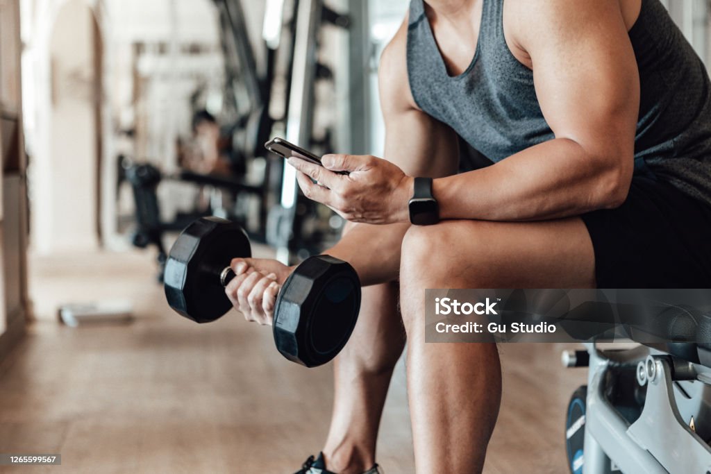 Muscular Young Man Training At Gym With Smart Phone Close up shot of a young man lifting dumbbell while using smartphone. Bodybuilding motivation and determination. Exercising Stock Photo