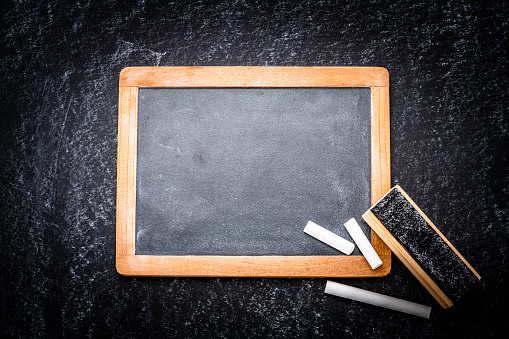 Top view of a blank blackboard with traces of chalk background with a board eraser and chalk placed at the bottom right corner of an horizontal frame leaving a useful copy space for text and/or logo. Predominant color is gray. High resolution 42Mp studio digital capture taken with Sony A7rII and Sony FE 90mm f2.8 macro G OSS lens