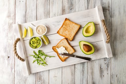 Toast with avocado, cream cheese, lime, guacamole and arugula on white wooden tray top view on wooden table.