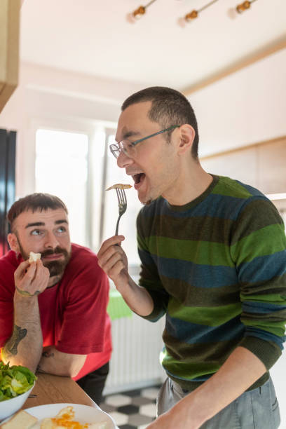 istock Man taking a bite with a fork, his boyfriend leaning on kitchen counter with a piece of bread 1265616713